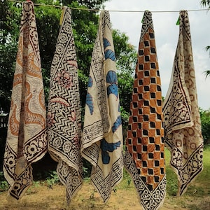 May include: Five patterned fabric scarves hanging on a clothesline. The scarves feature various designs in brown, black, and blue on a cream background. The patterns include floral, geometric, and abstract motifs. The scarves are hanging outdoors with a green tree background.