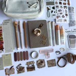 Puede incluir: Un conjunto de papelería que incluye un estuche beige, un cuaderno de tela marrón, varios bolígrafos, resaltadores y pegatinas. El set cuenta con clips con forma de café, un llavero de auriculares, rastreadores de hábitos y un bloc de notas.
