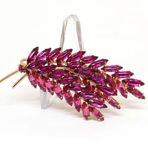 May include: A vintage brooch shaped like a leaf, with magenta and pink marquise-cut rhinestones set in gold-tone metal. The brooch is displayed on a clear stand against a white background.
