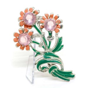 Puede incluir: Un broche de tono plateado con tres flores rosas con hojas y tallos verdes. Las flores tienen diamantes de imitación rosas en el centro.