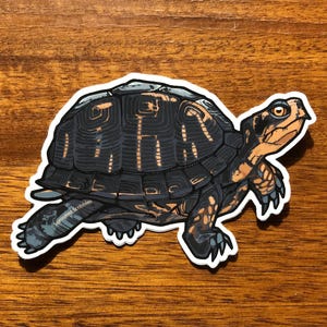 May include: A die-cut sticker featuring a detailed illustration of a turtle with a dark charcoal gray shell and orange-brown markings on its head, neck, and limbs. The text Sprouted Scribbles is printed in small white letters on the turtle's leg.