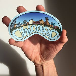 Op de afbeelding: Een blauwe en witte sticker met een stadsbeeld en het woord "Chicago" in een gestileerd lettertype.