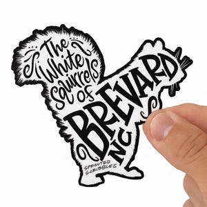 May include: A die-cut sticker shaped like a squirrel, featuring black text on a white background. The text reads The White Squirrels of BREVARD NC and SPROUTED SCRIBBLES. The design uses stylized lettering to form the silhouette of a squirrel.