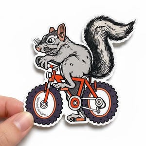 May include: A die-cut sticker featuring a gray squirrel with a large bushy tail riding a bright orange bicycle with thick black tires. The front tire has the text Sprouted Scribbles printed in small black letters along the inner rim.