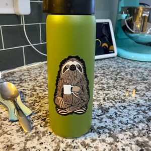 May include: A matte lime green cylindrical water bottle with a black lid. A sticker on the front depicts a brown, furry sloth sitting in a cross-legged position while holding a small white coffee mug. The sloth has dark eye markings.