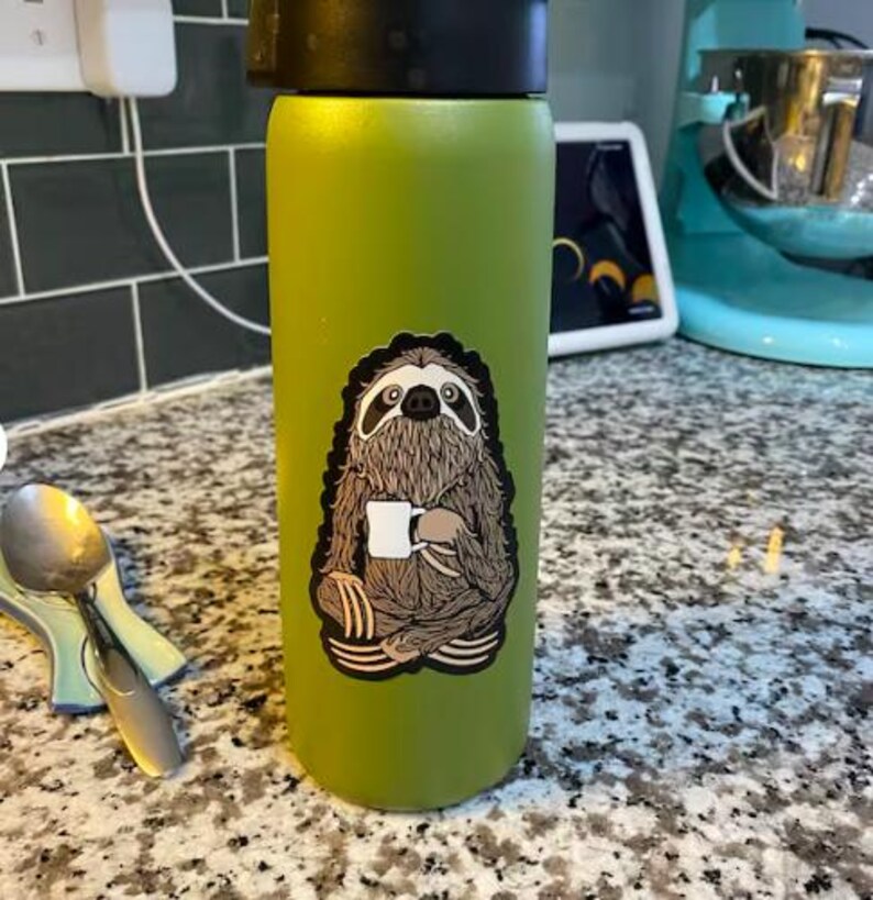 May include: A matte lime green cylindrical water bottle with a black lid. A sticker on the front depicts a brown, furry sloth sitting in a cross-legged position while holding a small white coffee mug. The sloth has dark eye markings.
