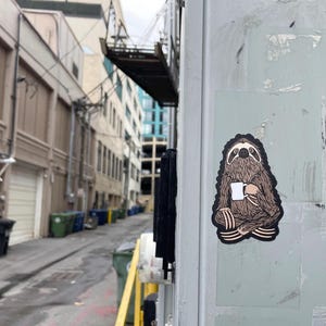 May include: A die-cut sticker featuring a brown, furry sloth sitting in a cross-legged position while holding a white mug. The sloth has dark brown fur details, a black outline, and a light-colored face. The sticker is adhered to a light gray surface.