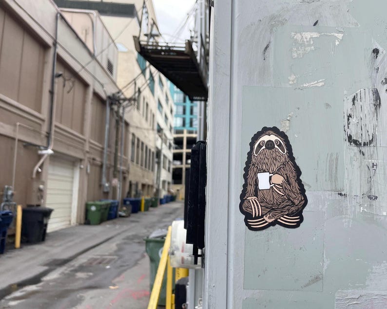 May include: A die-cut sticker featuring a brown, furry sloth sitting in a cross-legged position while holding a white mug. The sloth has dark brown fur details, a black outline, and a light-colored face. The sticker is adhered to a light gray surface.
