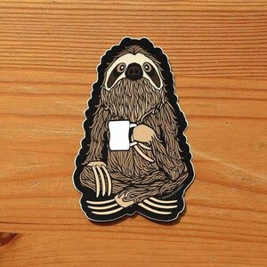 May include: A die-cut sticker featuring a brown, furry sloth sitting in a cross-legged position. The sloth has dark markings around its eyes and is holding a plain white mug with both hands. The illustration has a thick black border outline.