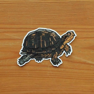 May include: A die-cut sticker featuring a detailed illustration of a turtle with a dark brown, textured shell and tan skin with black markings. The turtle is depicted in a side profile view, facing toward the right, with a white border outline.