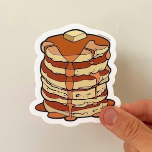 May include: A die-cut sticker featuring a stack of five golden-brown pancakes topped with a square pat of yellow butter and brown syrup dripping down the sides. The illustration includes the text Sprouted Scribbles in small black letters.
