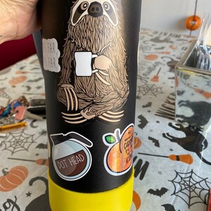 Puede incluir: Una botella de agua reutilizable negra con tapa amarilla y varias pegatinas. Las pegatinas muestran un perezoso sosteniendo una taza de caf&eacute;, una cafetera con el texto "Pot Head" y una naranja brillante con el texto "Chronic" e "Iconic".