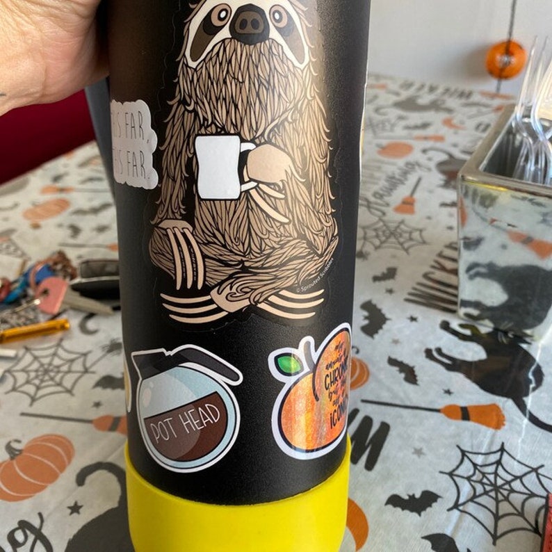 Puede incluir: Una botella de agua reutilizable negra con tapa amarilla y varias pegatinas. Las pegatinas muestran un perezoso sosteniendo una taza de caf&eacute;, una cafetera con el texto "Pot Head" y una naranja brillante con el texto "Chronic" e "Iconic".