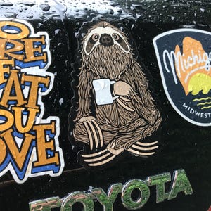 May include: A die-cut sticker featuring a brown sloth with detailed fur texture sitting in a cross-legged position while holding a small white coffee mug. The sloth has dark markings around its eyes and nose, and the sticker is applied to a dark surface.