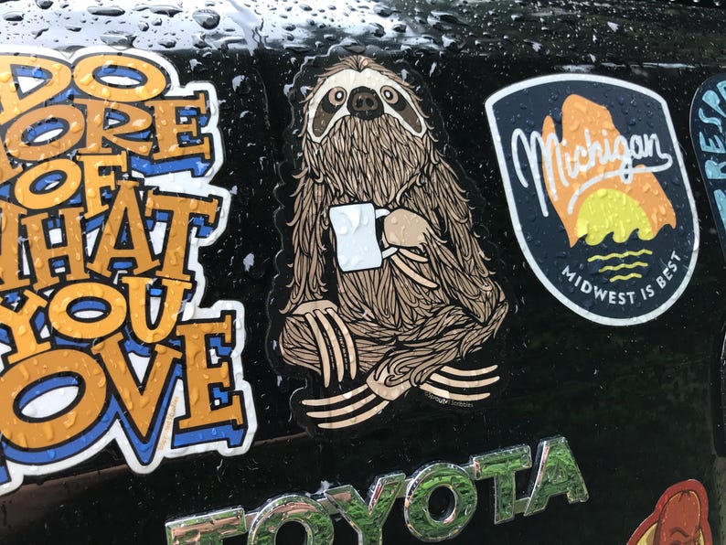 May include: A die-cut sticker featuring a brown sloth with detailed fur texture sitting in a cross-legged position while holding a small white coffee mug. The sloth has dark markings around its eyes and nose, and the sticker is applied to a dark surface.