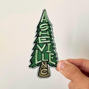 May include: An embroidered patch shaped like a pine tree with green foliage and a brown trunk. The text ASHEVILLE is stitched vertically in light green letters along the tree, with the letters NC stitched in brown on the trunk base.