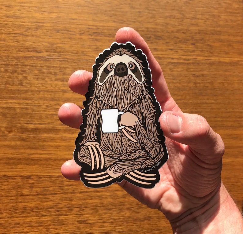 Puede incluir: Una pegatina de perezoso marr&oacute;n y negro con una taza blanca en su mano. La pegatina est&aacute; sobre una superficie de madera. El texto en la pegatina dice "Sprouted Scribbles".