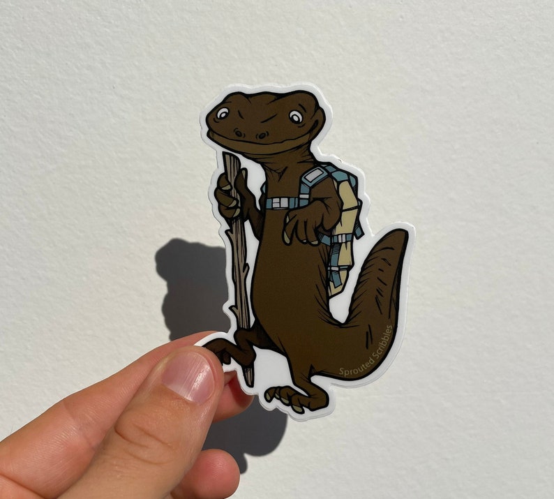Peut inclure: Un autocollant d&eacute;coup&eacute; repr&eacute;sentant un l&eacute;zard brun debout. Le l&eacute;zard porte un sac &agrave; dos bleu clair et beige et tient un b&acirc;ton de marche en bois. Le texte Sprouted Scribbles est imprim&eacute; en petites lettres blanches sur la queue du l&eacute;zard.
