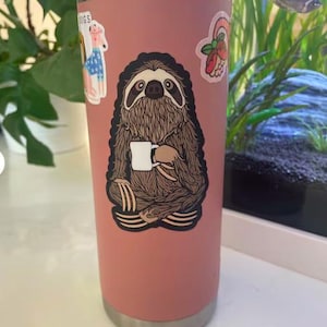 May include: A matte dusty rose cylindrical tumbler featuring a large central sticker of a brown sloth sitting in a cross-legged position while holding a small white mug. Additional stickers of a dog and a floral design are visible on the surface.