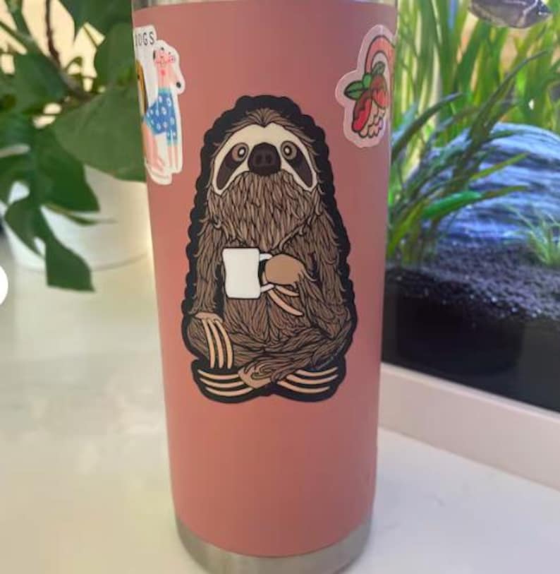 May include: A matte dusty rose cylindrical tumbler featuring a large central sticker of a brown sloth sitting in a cross-legged position while holding a small white mug. Additional stickers of a dog and a floral design are visible on the surface.