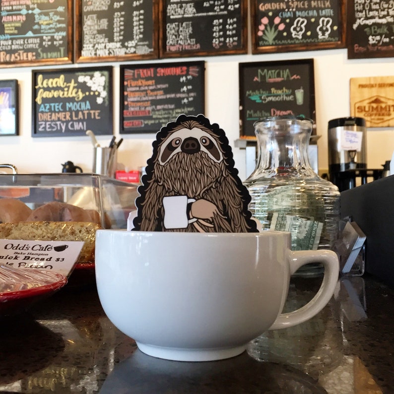 Puede incluir: Una taza de caf&eacute; de cer&aacute;mica blanca con una pegatina de perezoso en el lateral. El perezoso sostiene una taza de caf&eacute; y tiene una expresi&oacute;n relajada. La taza est&aacute; sobre una encimera de color marr&oacute;n oscuro.