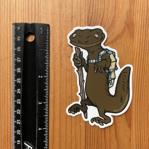 Peut inclure: Un autocollant repr&eacute;sentant une salamandre brune avec un sac &agrave; dos bleu clair et un b&acirc;ton de marche en bois. L'autocollant est plac&eacute; &agrave; c&ocirc;t&eacute; d'une r&egrave;gle noire indiquant une hauteur d'environ 10 cm. Le texte Sprouted Scribbles est imprim&eacute;.