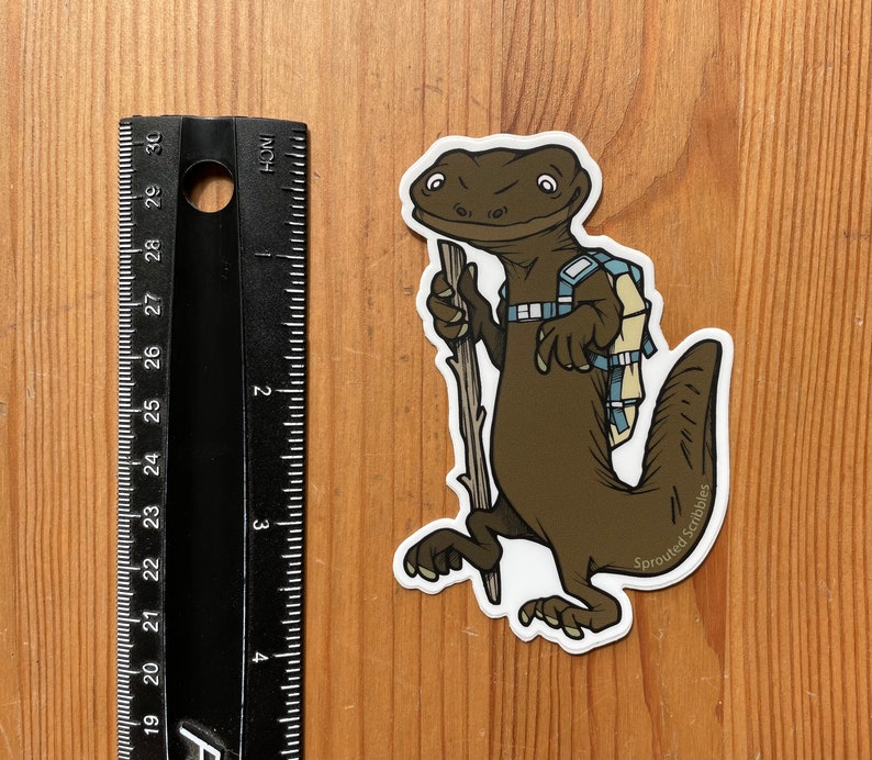 Peut inclure: Un autocollant repr&eacute;sentant une salamandre brune avec un sac &agrave; dos bleu clair et un b&acirc;ton de marche en bois. L'autocollant est plac&eacute; &agrave; c&ocirc;t&eacute; d'une r&egrave;gle noire indiquant une hauteur d'environ 10 cm. Le texte Sprouted Scribbles est imprim&eacute;.