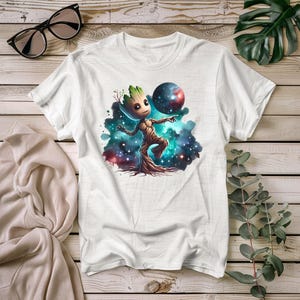 May include: White t-shirt featuring a cartoon character resembling a tree, holding a planet-like sphere. The design includes a colorful nebula background with stars. The shirt is displayed on a wooden surface with sunglasses and a folded cloth.