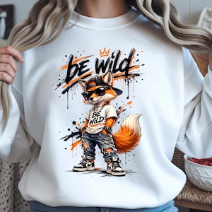 Può includere: Felpa bianca con un personaggio di una volpe dei cartoni animati con occhiali da sole, berretto e jeans. La volpe ha la pelliccia arancione e bianca e si trova di fronte a schizzi di vernice nera e arancione. Le parole "BE WILD" sono stampate.