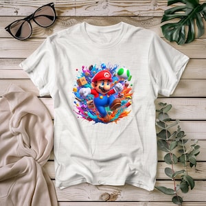 Könnte beinhalten: Weißes T-Shirt mit einer farbenfrohen Grafik von Mario aus dem Videospiel Super Mario Bros. Mario ist in seinem klassischen roten Hut und blauen Overalls dargestellt, umgeben von lebendigen Farbspritzern und Spielelementen.