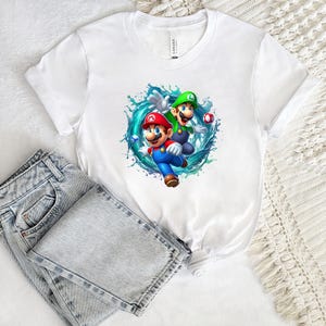 Può includere: T-shirt bianca con una grafica colorata di Mario e Luigi circondati da spruzzi d'acqua. I personaggi indossano i loro abiti classici. Sono visibili anche jeans di denim color azzurro chiaro.