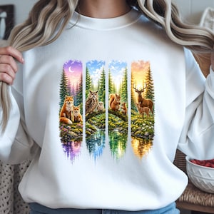 Op de afbeelding: Witte sweatshirt met een kleurrijke natuurscène met een vos, uil, eekhoorn en hert. Het ontwerp omvat een bos, waterreflectie en een zonsondergang/zonsopgang achtergrond, verdeeld in verticale panelen.