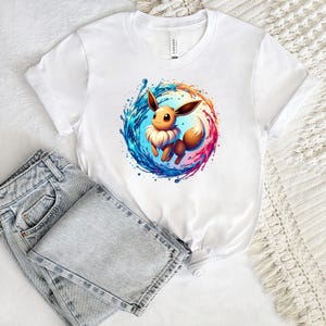 May include: White t-shirt with a cartoon character graphic, encircled by blue, pink, and orange splashes. The shirt is styled with light-wash denim jeans. The top has a round neck and short sleeves. The image shows a casual outfit.