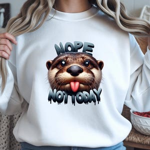 Puede incluir: Sudadera blanca con una ilustración detallada de una nutria sacando la lengua. Las palabras "NOPE" y "NOT TODAY" están impresas encima y debajo de la nutria en una fuente 3D.