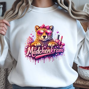 Peut inclure: Sweat-shirt blanc avec un guépard portant des lunettes de soleil roses et un nœud, avec le texte allemand "Mädchenkram" en rose et violet. Le motif comprend des cupcakes, des cœurs et du maquillage.