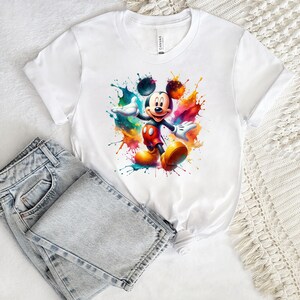 Könnte beinhalten: Weißes T-Shirt mit einem farbenfrohen Aquarelldesign von Mickey Mouse. Die Zeichentrickfigur ist von lebendigen Farbspritzern in Blau-, Orange- und Gelbtönen umgeben. Das Shirt wird mit hellblauen Jeans kombiniert.