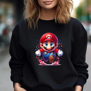 Può includere: Felpa nera con una grafica colorata del personaggio del videogioco Mario. Il personaggio dei cartoni animati è raffigurato con il suo iconico cappello rosso, la tuta blu e i guanti bianchi, che corre con un'espressione determinata. Il design include accenti colorati.