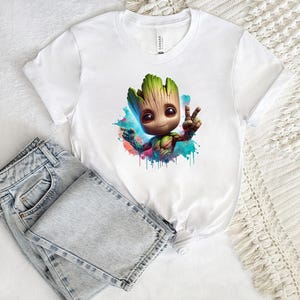 May include: White t-shirt featuring a colorful illustration of a cartoon character giving a peace sign. The character has a wooden appearance with green and brown tones, set against a watercolor splash of blue, pink, and green. Light-wash denim jeans are folded next to the shirt.