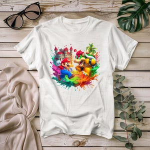 May include: White t-shirt with a colourful graphic of Mario and Bowser from the Super Mario video game series. The design includes a castle, vibrant paint splashes, and the characters in action. The shirt is made of a soft, comfortable material.