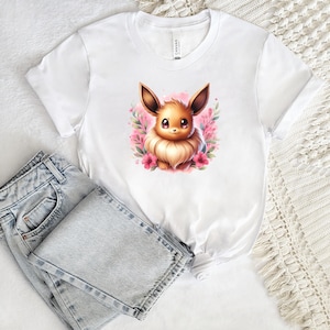 May include: White t-shirt featuring a watercolor illustration of a cartoon Eevee character surrounded by pink flowers. The shirt is paired with light-wash denim jeans. The shirt is made of a soft material.