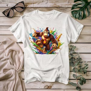 May include: White t-shirt featuring a colorful graphic of Donkey Kong with a red and gold medallion. The cartoon ape is depicted in a dynamic pose against a backdrop of vibrant paint splashes. The shirt is displayed on a wooden surface.