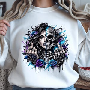 May include: White sweatshirt with a striking graphic of a woman's face split with a skull. The design incorporates blue and purple roses, and the middle finger is raised. The artwork has a tattoo-style aesthetic with a splash of blue and purple paint.