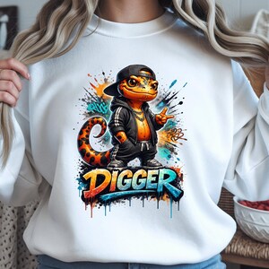 Peut inclure: Sweat-shirt blanc avec un lézard de dessin animé portant une casquette et une veste noires, avec le mot "DIGGER" dans un style graffiti coloré. Le lézard est orange avec une queue tachetée, faisant un signe de paix.