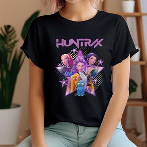 Könnte beinhalten: Schwarzes T-Shirt mit einer Grafik von drei Frauen und einer blauen Kreatur in einem Stern, mit dem Wort "HUNTRAX" in Lila. Das Design hat einen futuristischen, animierten Stil. Das T-Shirt wird von einer Person getragen.