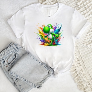 May include: White t-shirt with a colourful graphic of a green cartoon dinosaur character and a splash of rainbow paint. The shirt is paired with light wash denim jeans.