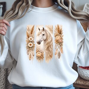 Op de afbeelding: Witte sweatshirt met een aquarelontwerp van een wit paard. Het paard wordt geflankeerd door drie verticale panelen met bloemen- en veermotieven in beige en bruin. Het ontwerp is in een zachte, artistieke stijl, geschikt voor vrijetijdskleding.