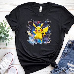 May include: Black t-shirt with a colourful graphic of Pikachu, a yellow Pokémon character, set against a cosmic background. The design includes shades of blue, purple, and yellow. The shirt is paired with blue jeans and white trainers.