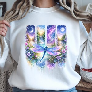 Peut inclure: Sweat-shirt blanc orné d'un motif graphique coloré de style aquarelle. Le design présente une libellule aux ailes violettes et bleues, sur fond de quatre panneaux verticaux représentant des paysages avec des lunes, des cascades et des ciels étoilés. Des motifs floraux encadrent les panneaux.