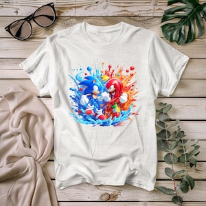 May include: White t-shirt with a colourful graphic of Sonic the Hedgehog and Knuckles, set against blue and orange splashes. The design shows the characters in action, with vivid colours and detailed artwork. The shirt is made of a soft material.