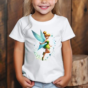 Può includere: T-shirt bianco con una fata dei cartoni animati con capelli biondi, vestito verde e ali blu. La fata è circondata da elementi in stile acquerello blu e verdi. La maglietta è indossata da un bambino.